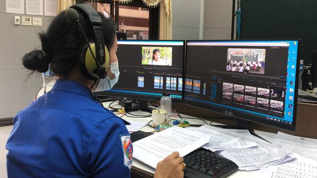 A woman wearing headphones sits at a desk with her back to the camera, working on a dual screen computer